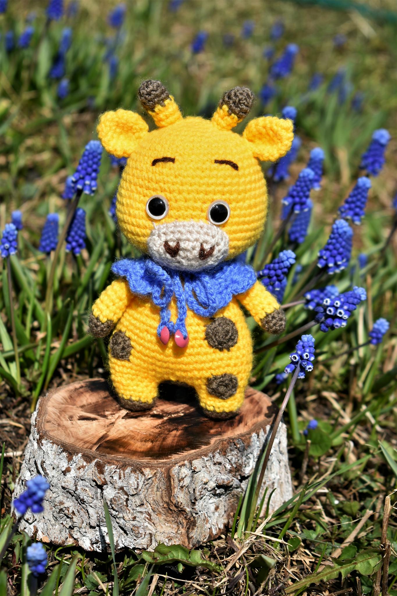 Cute crocheted giraffe toy surrounded by vibrant purple flowers on a sunny day.